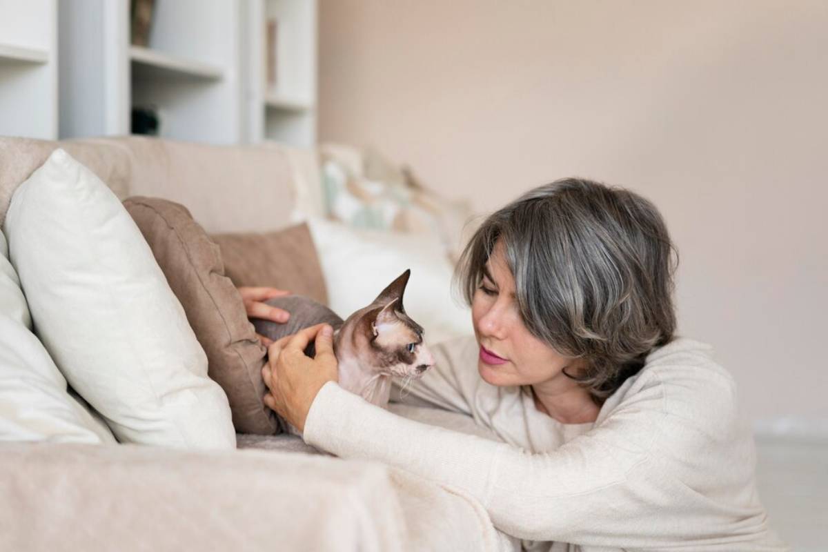 Middle-aged woman in a beige sweater lovingly looking at her Sphynx cat on a couch.