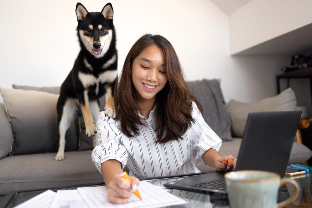 Young woman working on laptop with a black and tan dog standing on the couch behind her.