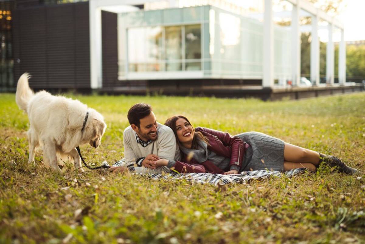 A joyful couple lies on a blanket in a park, smiling and watching their dog nearby.
