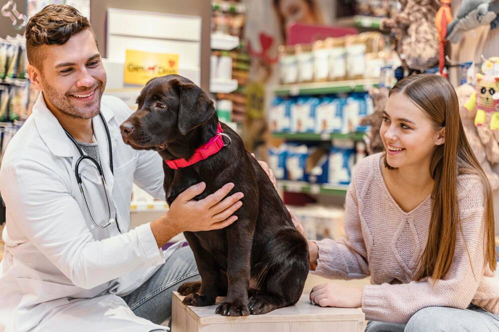 A veterinarian and a young woman smile at a black dog in a pet store.