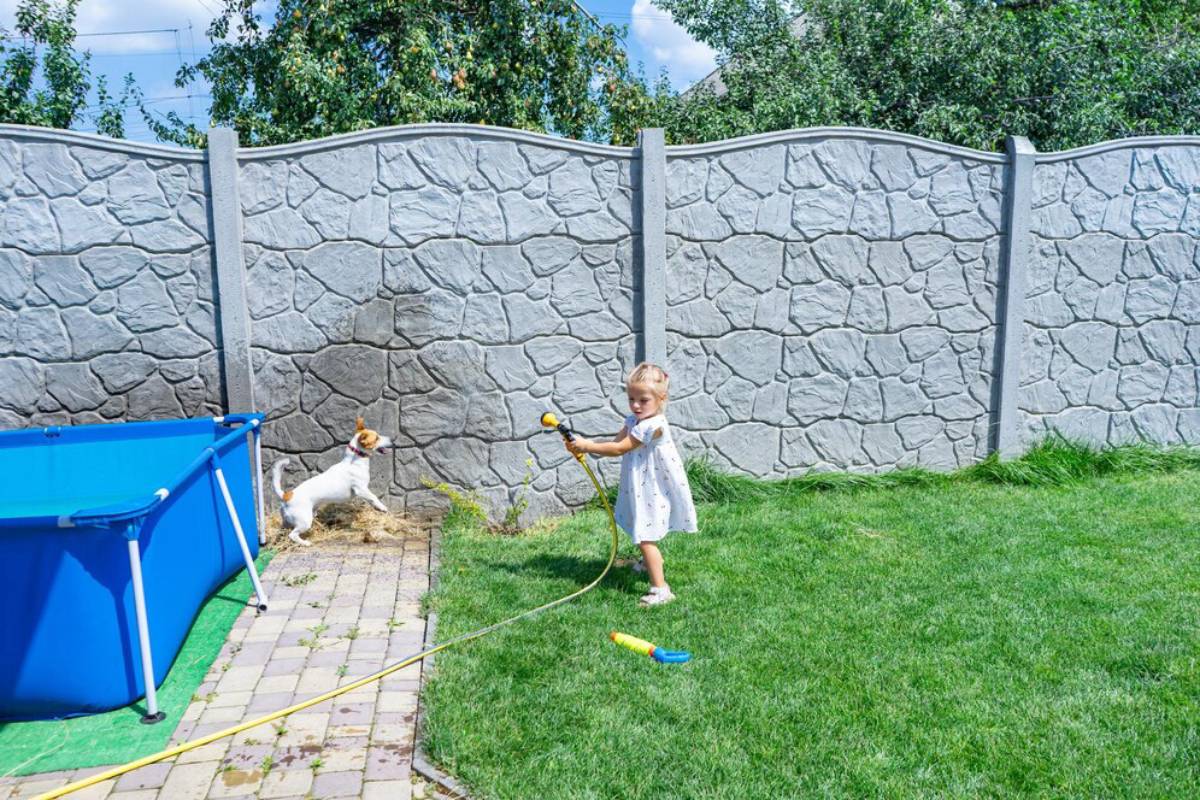 Toddler playing with a hose in a sunny backyard with a dog jumping near a blue pool.