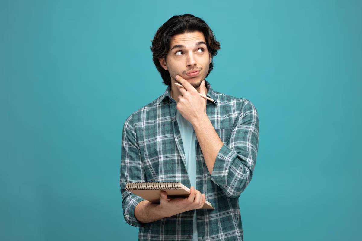 Young man in a plaid shirt holding a notebook and looking thoughtful, against a teal background.