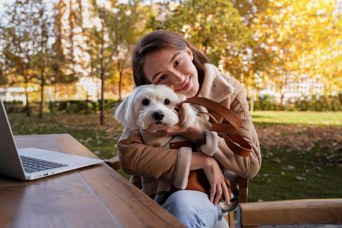 Young woman and her small white dog sitting at a park table near a laptop in autumn.