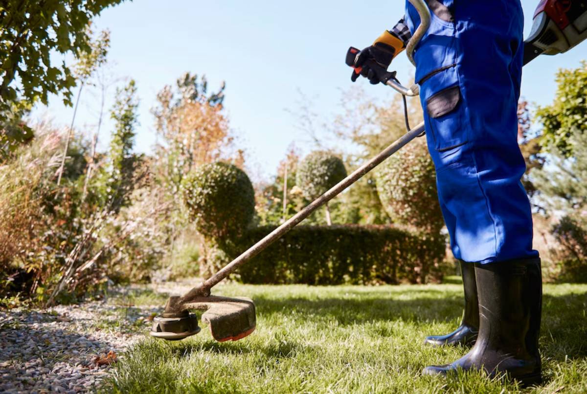 Person in blue coveralls using a weed trimmer in a sunny garden.