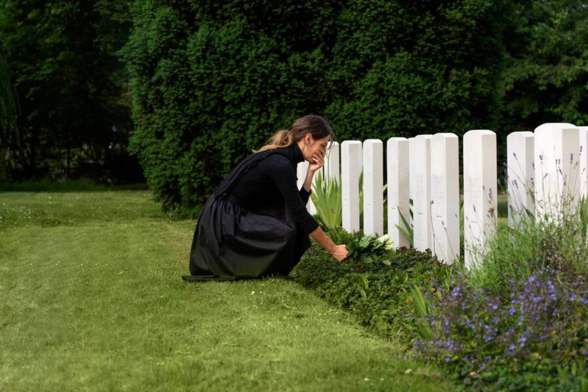 A man in a suit places a rose on a stone memorial in a grassy area.