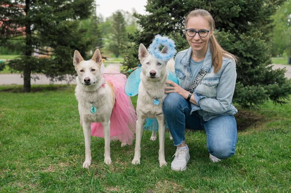 Woman in glasses and denim jacket kneeling next to two decorated dogs in a park.