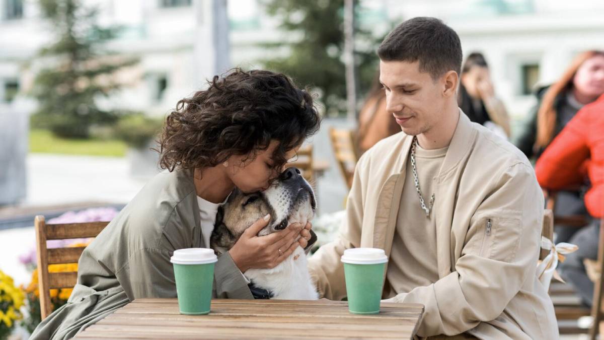 A woman affectionately kisses a dog on the head while sitting at an outdoor café with a man.