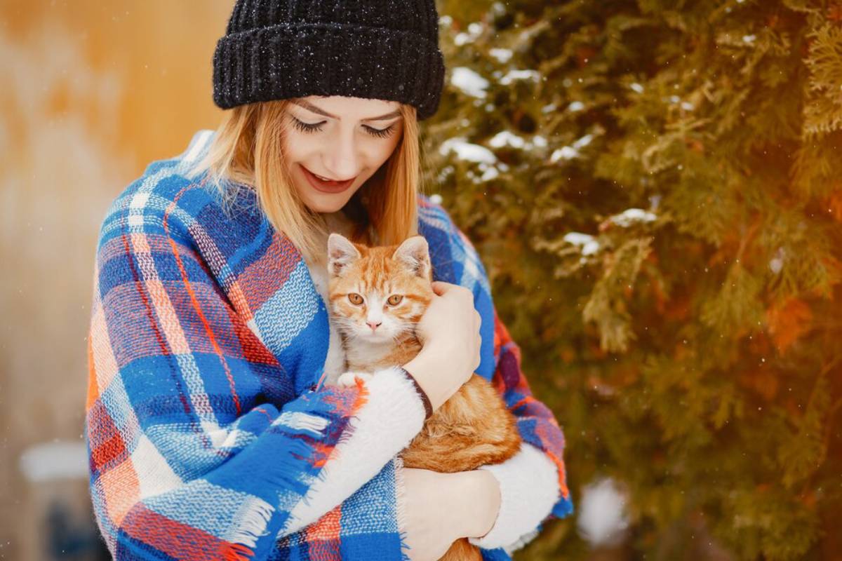 A woman in a winter hat and colorful scarf holds an orange cat, smiling, in a snowy setting.